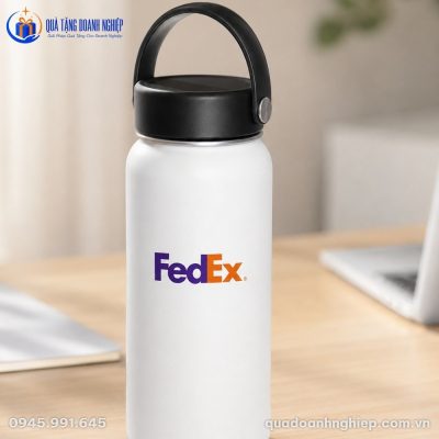 Cận cảnh bình giữ nhiệt in logo FEDEX màu trắng nhám đặt trên bàn làm việc gỗ cạnh máy tính