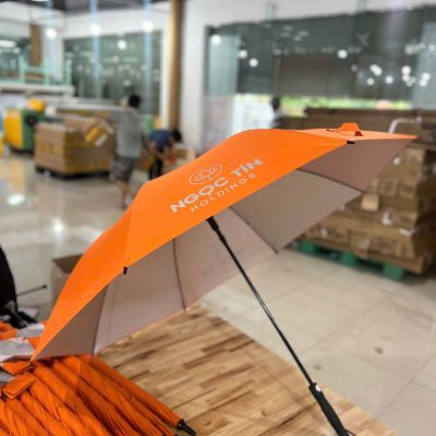 Góc nghiêng của ô dù màu cam in logo trắng nổi bật trên nền bàn gỗ công nghiệp