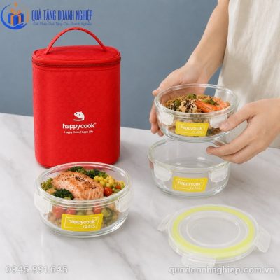 Người dùng đang cầm một hộp thủy tinh tròn đựng mì xào tôm, bên dưới là một hộp rỗng và một hộp khác đựng cá hồi áp chảo kèm rau củ kế bên túi giữ nhiệt màu đỏ