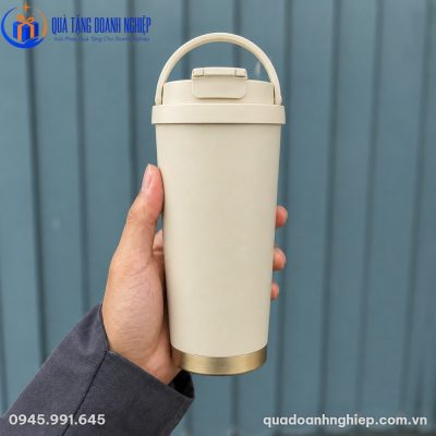 Cận cảnh tay người dùng cầm chiếc ly giữ nhiệt màu be nhám mờ với quai xách tiện lợi trên nền hàng rào màu xám xanh