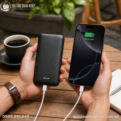 Người dùng cầm trên tay sạc dự phòng Akus và điện thoại đang sạc trong không gian quán cà phê sân vườn