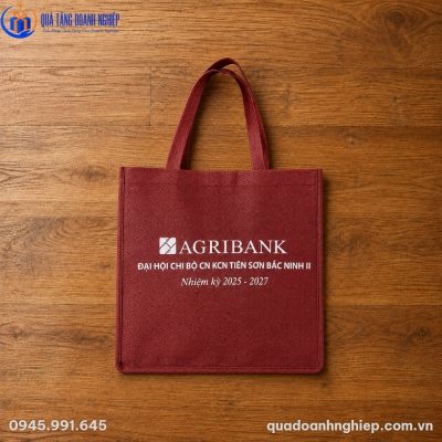 Túi vải không dệt màu đỏ đô Agribank đặt phẳng trên nền sàn gỗ màu nâu ấm sang trọng, làm nổi bật logo in lụa sắc nét