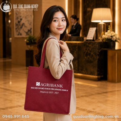 Nữ nhân viên công sở Việt Nam thanh lịch trong trang phục vest màu kem đang đeo chiếc túi vải Agribank màu đỏ đô trên vai tại sảnh khách sạn cao cấp
