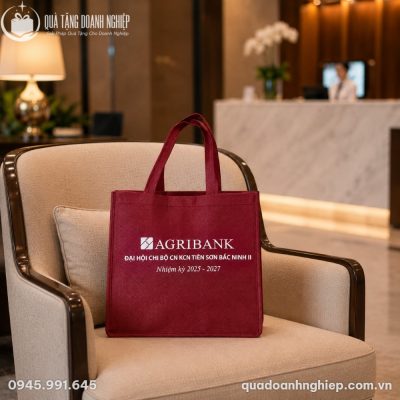 Chiếc túi vải không dệt màu đỏ đô Agribank được đặt trang trọng trên chiếc ghế bành bọc nệm màu kem trong không gian phòng chờ sang trọng của khách sạn
