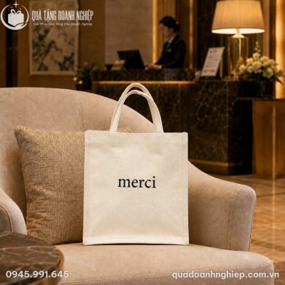 Túi canvas in chữ Merci đặt ngay ngắn trên ghế bành da trong phòng chờ VIP